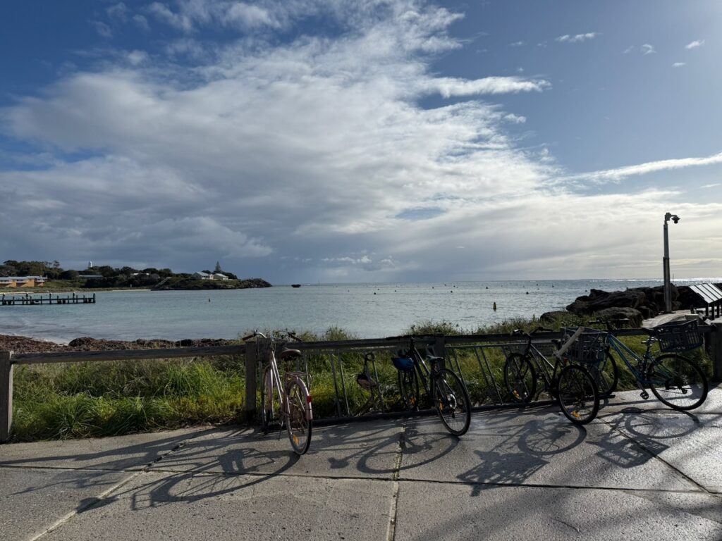 A bike rack on the ferry jerry looking across Thomson Bay, which is calm and sunlit, though there are growing clouds. Barely visible in the distance is the island's second lighthouse, Bathurst Lighthouse, on Bathurst Point.