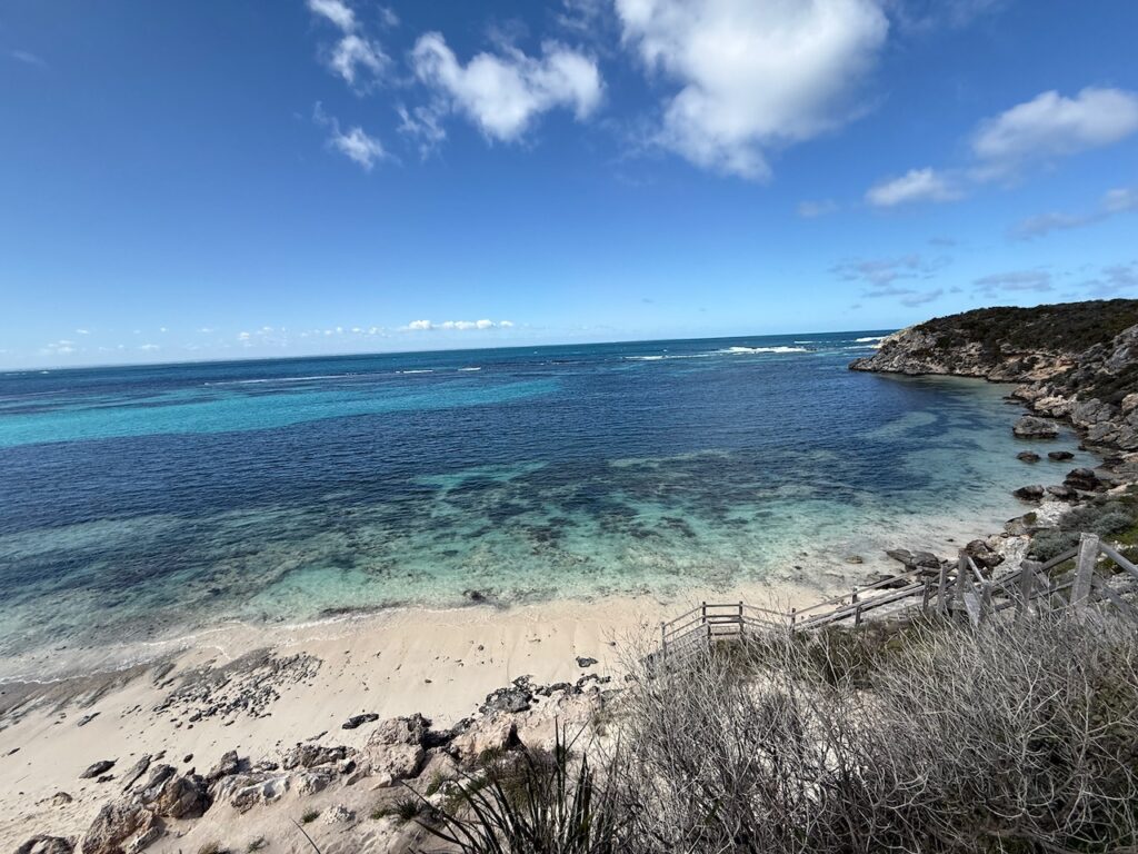 Wooden stairs on vegetated limestone lead down to sandy beach and a calm bay showing large areas of coral under the water
