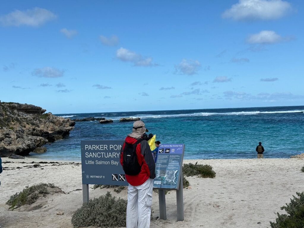 Doug, wearing a red coat, hat and backpack, reads an information sign in front of Little Salmon Bay at Parker Point
