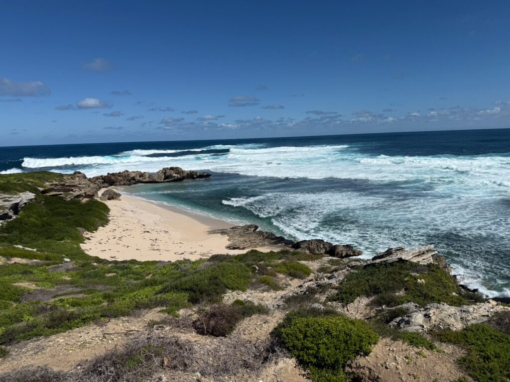 Waves on blue ocean, rocks jutting into the sea, forming a small sandy cove, edged by green vegetation