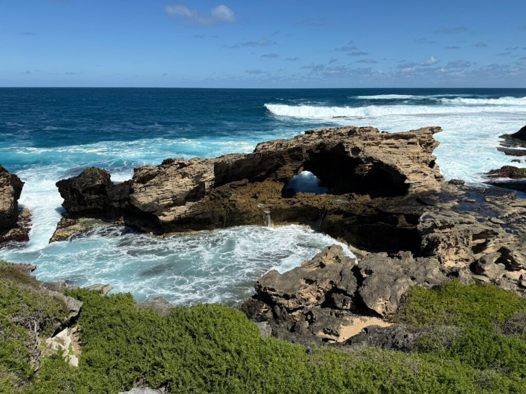 Eroded limestone outcrop making a small bridge, among waves on a deep blue ocean and a blue sky. Green vegetation covers the foreshore