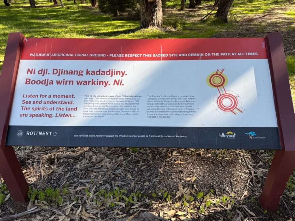 Red white and green sign showing Indigenous art and information about the Indigenous burial ground at the Settlement. It reads: "WADJEMUP ABORIGINAL BURIAL GROUND • PLEASE RESPECT THIS SACRED SITE AND REMAIN ON THE PATH AT ALL TIMES Ni dji. Djinang kadadjiny. Boodja wirn warkiny. Ni. Listen for a moment. See and understand. The spirits of the land are speaking. Listen... Here lie the unmarked graves of over 370 Aboriginal men and boys. Their stories are forever linked to this land, yet they rest a long way from home. Between 1838 and 1931 Wadjemup was a prison for at least 3700 Aboriginal men and boys from across Western Australia. Leaders, law men, warriors, fathers, brothers and sons were forced to leave their communities and families. Those who died on Wadjemup remain here amongst the trees. Their spirits are part of the island. The Whadjuk Traditional Owners, representative Aboriginal Groups from across the state, Rottnest Island Authority, Wadjemup Aboriginal Reference Group, Rottest Foundation and other partners are working towards a culturally appropriate .. acknowledgement of this site's history. This is a journey towards reconciliation and healing for the physical and spiritual restoration of this place. The past is not buried. ROTTNEST IS The Rottnest Island Authority respect the Whadjuk Noongar people as Traditional Custodians of Wadjemup"