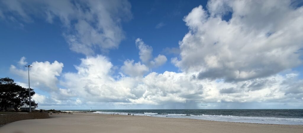 City Beach, Perth. Long stretch of sand, waves, and a deep green ocean under a blue sky with many clouds, In the distance, heavy rain is approaching