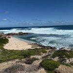 Waves on blue ocean, rocks jutting into the sea, forming a small sandy cove, edged by green vegetation
