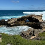 Eroded limestone outcrop making a small bridge, among waves on a deep blue ocean and a blue sky. Green vegetation covers the foreshore
